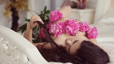 A person with light white skin and long brown, wavy hair reclines on a white bed in a sensual pose, holding pink flowers with long green stems over their chest and body with the right hand, and with the left hand pulling gently on a trellis of hair. This image represents the aspects of sensuality, sexuality, and masturbation that I include in my dilator journey.