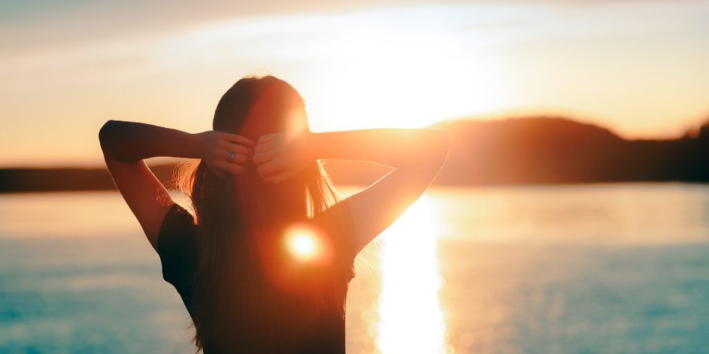 The back of a person from the ribcage up with their hands resting loosely on the back of their head with elbows out to either side,  looking out into a beautiful sunset that reflects brightly off the expanse of water before them. This image represents the concept of hope with Lichen Sclerosus.