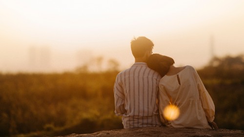 Image of two people from behind sitting in front of a vast open landscape of trees and grass. The person on the right wears a loose-fitting light colored blouse and bends their neck to lean their head on the other person's shoulder, who is sitting upright in a white colored shirt with vertical dark stripes. The sun is setting in the distance, providing a warm sepia appearance. This image represents the intimacy and vulnerability my husband and I experienced while working with dilators.