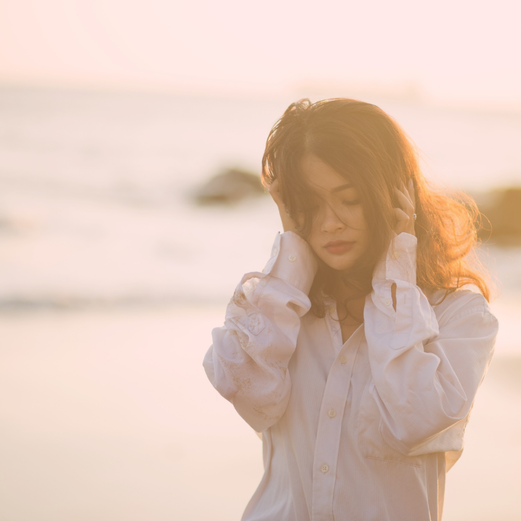 A person with light skin and long, tousled dark hair walks along a beach with eyes cast down and hair partially covering one eye. They are wearing a button-down collared long sleeved shirt and gingerly holding both hands over either side of their head. The image’s purpose is to represent someone feeling sad and hopeless.