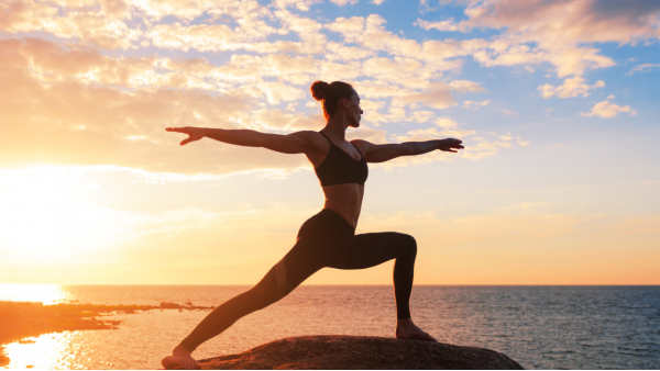 Image of a person doing a warrior 2 yoga pose, outside, on top of a rock. They're wearing a black bralette and black full-length yoga pants and have a tight bun hairstyle. The background is a beautiful sunrise with blue, orange, and yellow skies and soft, fluffy white clouds above a calm ocean.