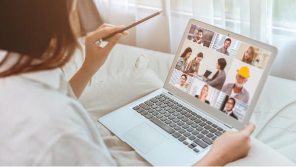 Over the right shoulder view of someone seated on a white-linen bed, with a laptop resting on a pillow. They have brown shoulder length hair and are wearing a white collared shirt. On the laptop screen is a virtual meeting with a matrix of nine separate meeting windows, one for each attendee. The attendee images are blurry. The person loosely holds a pencil in their left hand and is pointing it just above the screen. The image represents the Lichen Sclerosus Support Virtual Meetups.