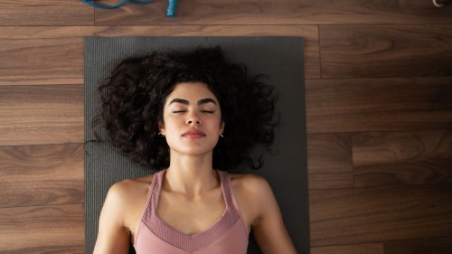 Image of a person with dark brown hair and brown skin lying on top of a dark grey yoga matt, on a wooden yoga studio floor. The person is wearing a dusty rose tank top and their eyes are closed with a peaceful look on their face.